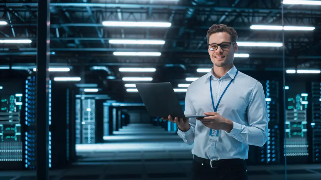 Smiling IT professional holding a laptop in a modern data center. The background features rows of server racks illuminated by blue lights, creating a high-tech environment. The individual is wearing a light blue shirt and a lanyard, representing expertise in data management and IT infrastructure.