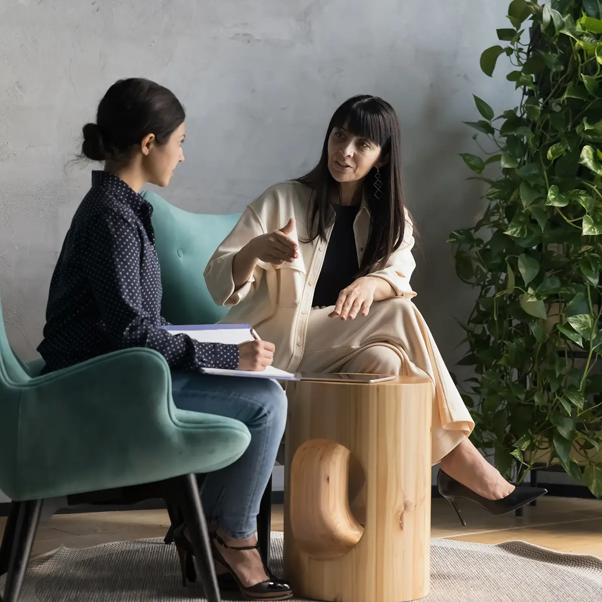 Two women are engaged in a mentoring session. One woman, dressed in a light-coloured outfit, is speaking animatedly, using hand gestures to emphasise her points. She sits in a modern, comfortable chair beside a stylish wooden side table. The other woman, wearing a dark blouse and jeans, listens attentively while taking notes on a pad of paper. The setting is cosy and modern, with a potted plant adding a touch of greenery to the scene.