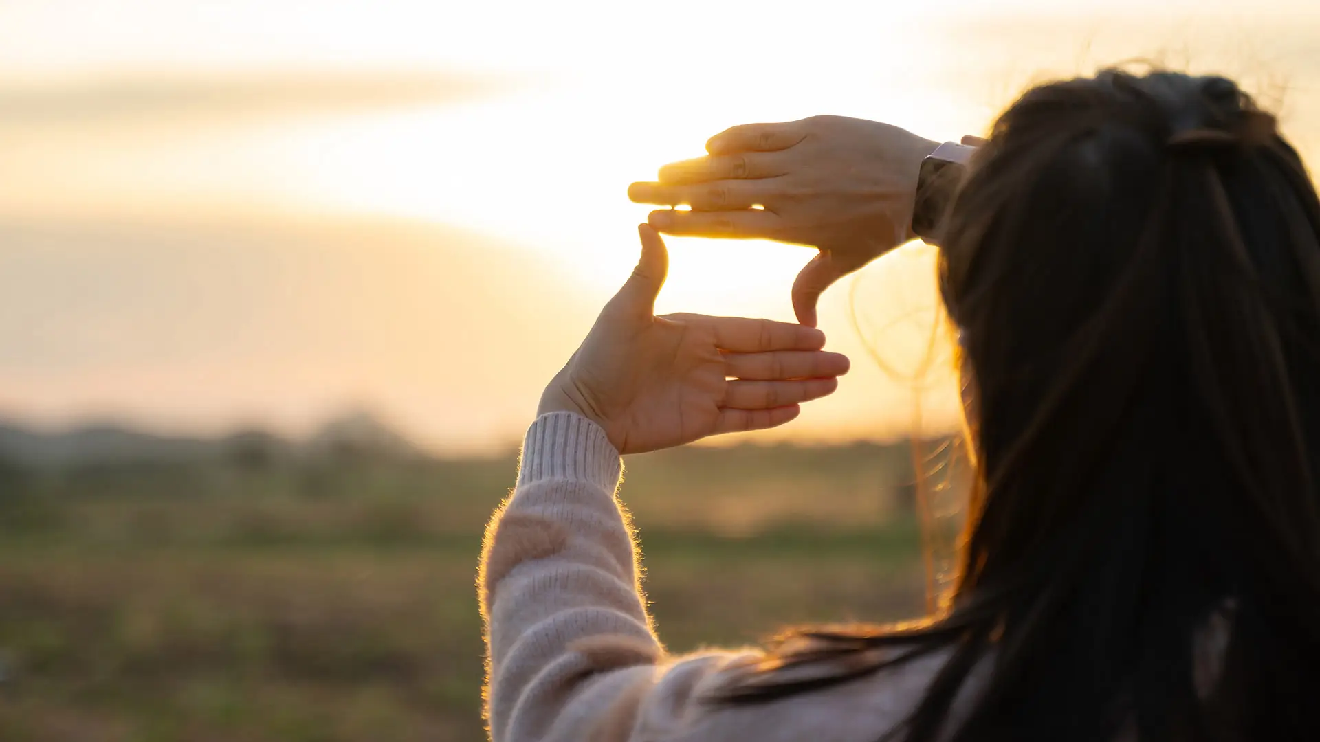 A woman with long dark hair and a light sweater stands outdoors at sunset, forming a rectangular frame with her hands as she looks towards the sun. The warm golden light creates a silhouette effect, symbolising vision, focus, and new beginnings.