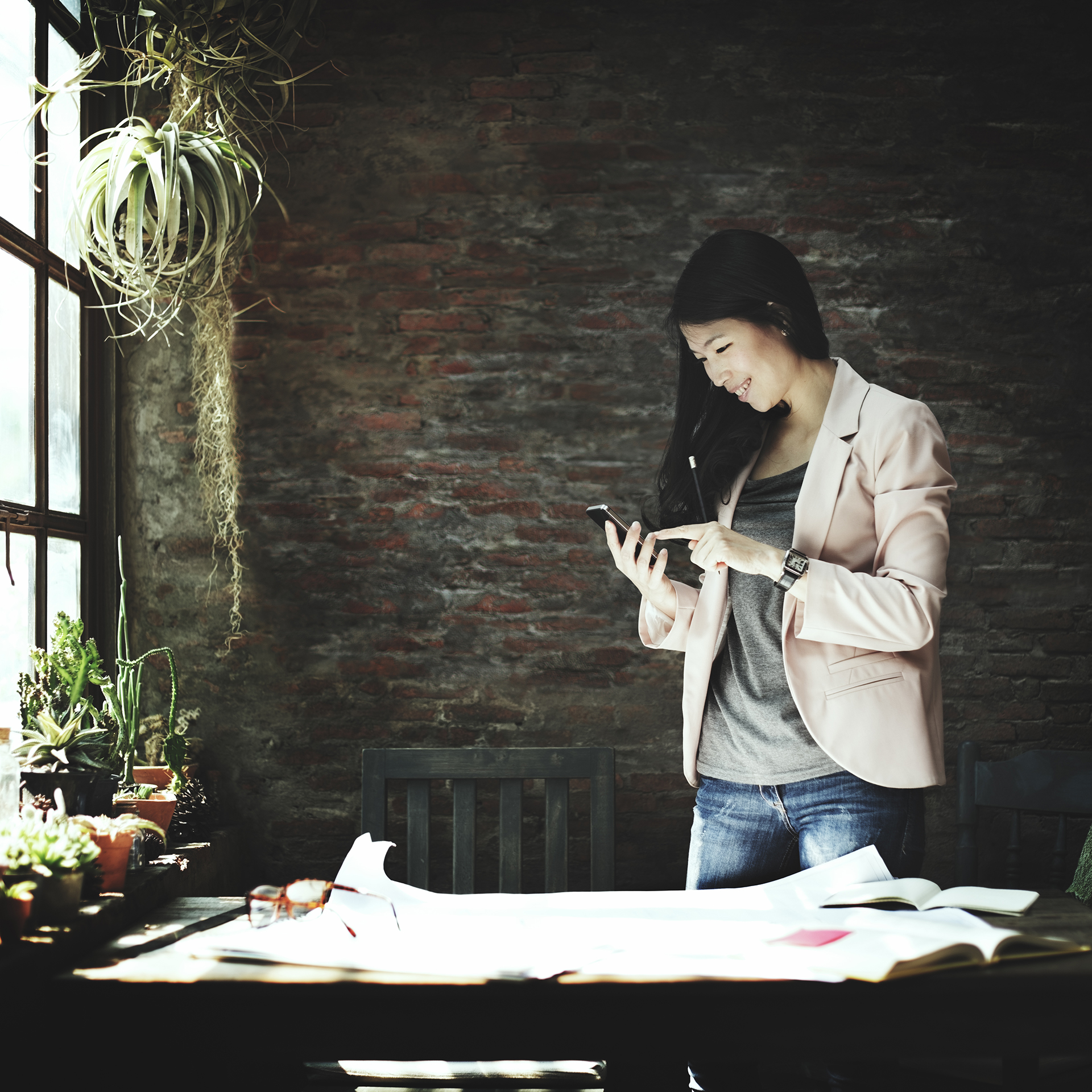 The image features a young professional woman standing by a desk in a well-lit room with a rustic brick wall. She is smiling and looking at her phone, dressed in a casual yet professional outfit with a blazer, jeans, and a t-shirt. The desk is scattered with open books and papers, suggesting a workspace. This image is perfect for representing a "Bachelor of Business" program, highlighting a modern and dynamic approach to business education.