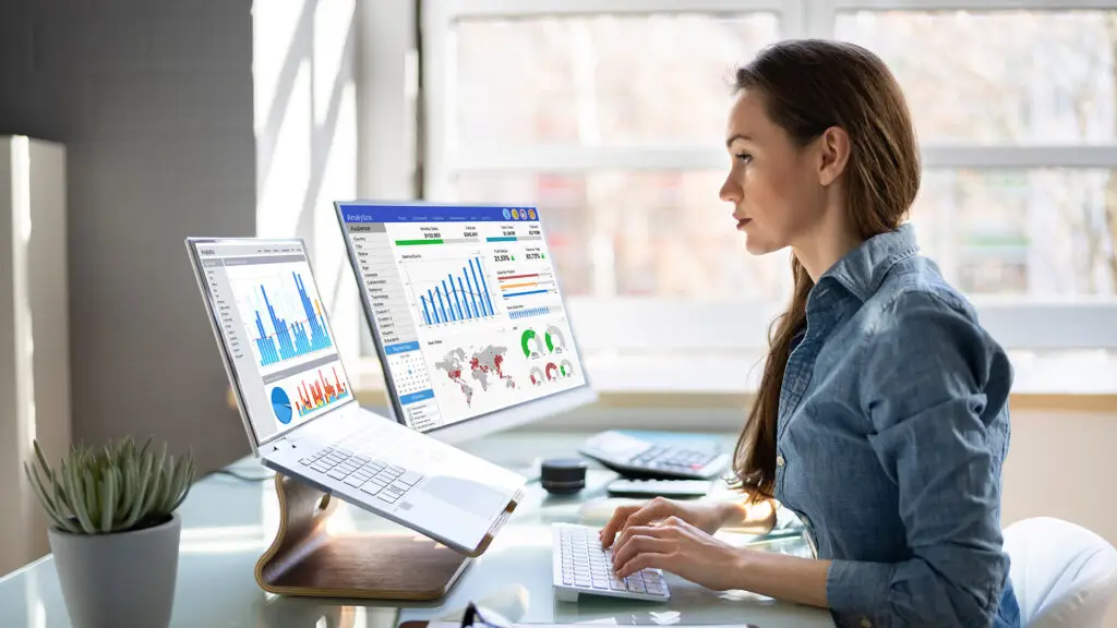 Business analyst working at a desk with dual monitors displaying various analytics dashboards, charts, and graphs. The individual is focused on reviewing data, with a laptop and external keyboard in front of them. The office setting is bright and modern, with natural light coming through large windows.