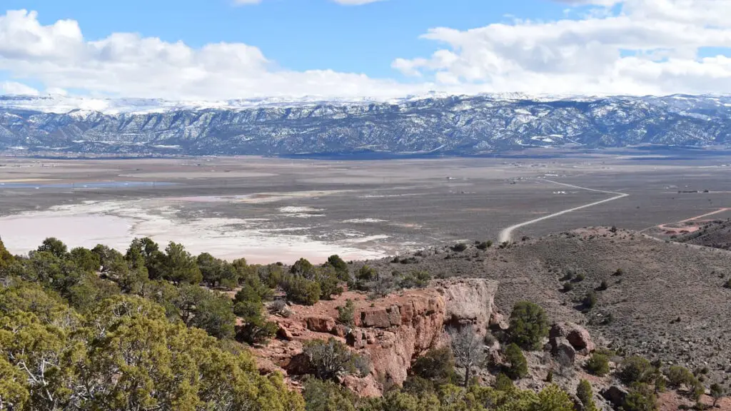 A scenic view of the Cedar Mountains in Utah, showcasing a vast, open landscape with a mix of rugged terrain and sparse vegetation. Snow-capped peaks rise majestically in the background under a bright blue sky with scattered clouds, while a winding road cuts through the expansive valley below, leading the viewer's eye through the serene and untouched beauty of the region.