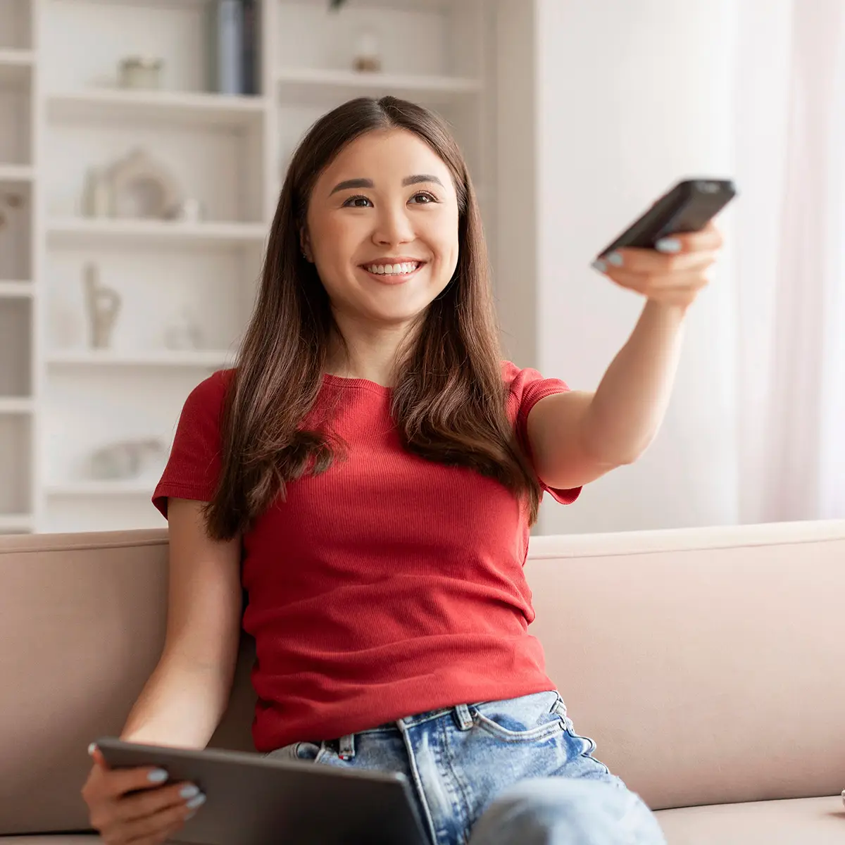 A person sits comfortably on a couch, smiling while holding a remote control in one hand and a tablet in the other, representing the concept of flexible learning on your own terms. The relaxed, home environment with shelves in the background suggests the freedom to manage study and leisure time effectively.