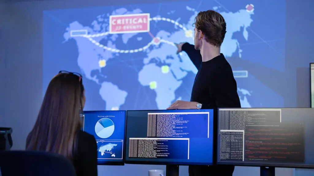 Cybersecurity professionals monitoring critical global events on a large screen map. One individual points to a location on the map indicating a 'Critical Events' alert, while another observes data on multiple monitors displaying vulnerabilities and system logs. The room has a high-tech and security-focused atmosphere.