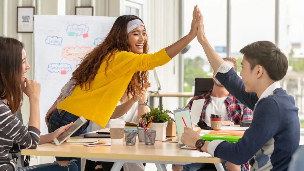 A diverse group of young adults is enthusiastically engaged in a collaborative session. They are gathered around a table, equipped with tablets, notebooks, and coffee cups, indicating a productive and dynamic environment. The scene captures a moment of high energy and camaraderie as a young woman in a bright yellow sweater high-fives a fellow participant. The background features a whiteboard filled with colorful diagrams and keywords such as "strategy," "innovation," and "leadership," highlighting a brainstorming or planning activity. This vibrant and inclusive setting embodies the spirit of equal access to education and shared success, emphasizing teamwork and mutual support among peers.