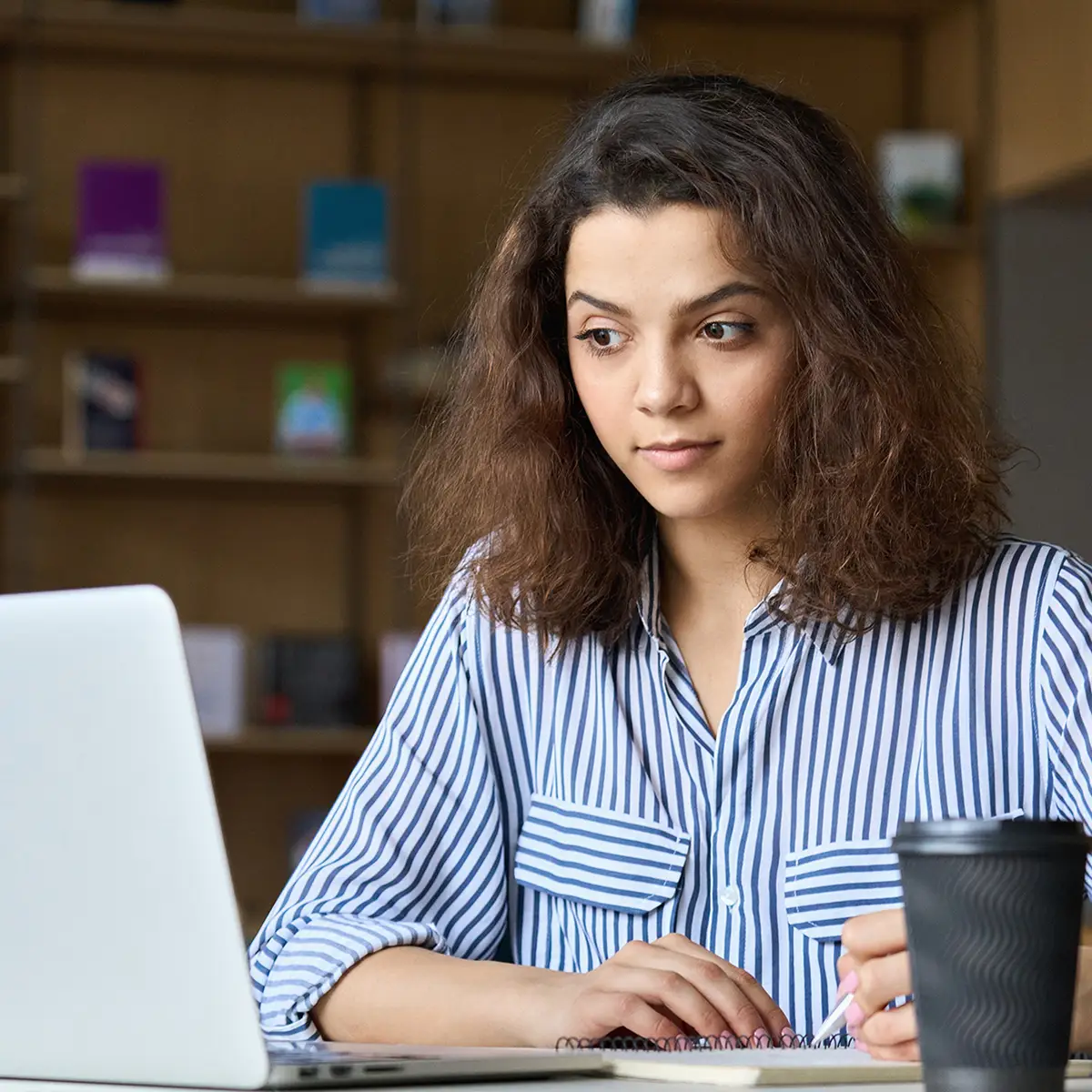 A young woman with curly hair sits at a desk in a library or study area, focused intently on her laptop screen. She wears a striped shirt and has a notebook open in front of her, with a pen in her hand, suggesting she is taking notes. A takeaway coffee cup is placed nearby. The background shows blurred bookshelves, enhancing the academic environment.