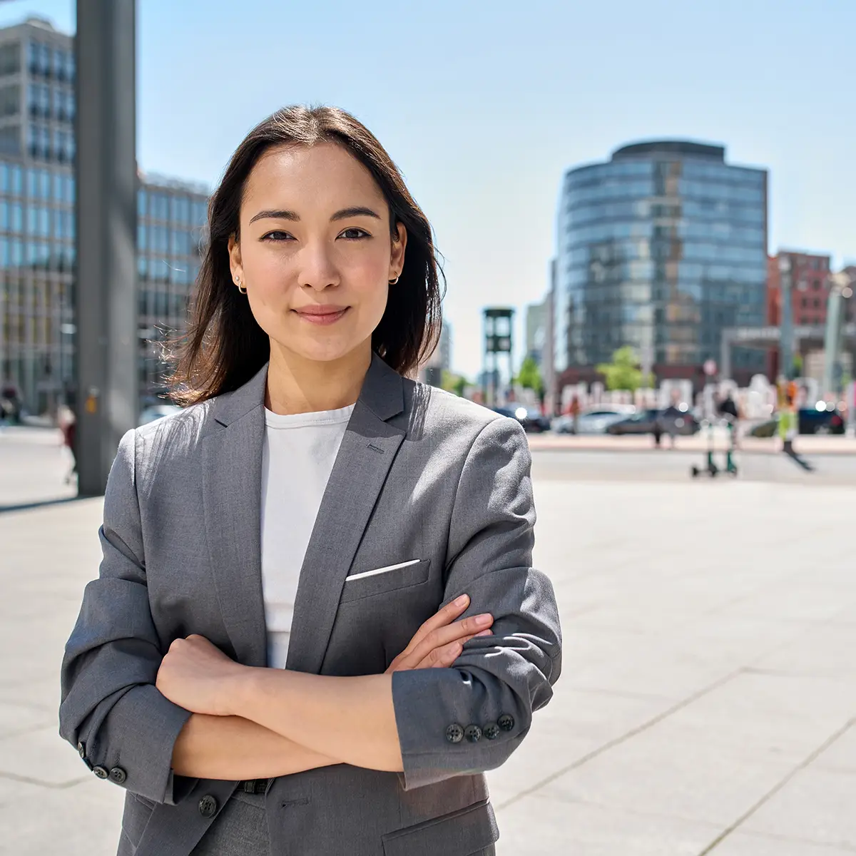 A confident young businesswoman stands outdoors in an urban setting, with modern office buildings in the background. She is wearing a grey suit and white shirt, with her arms crossed and a determined expression on her face. The bright daylight and clear sky suggest a positive and dynamic environment.