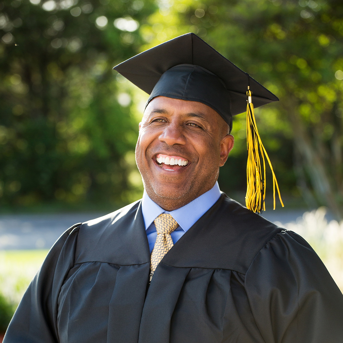 The image shows a mature man in graduation attire, smiling broadly, with a background of greenery and sunlight. This image can represent "Happy graduate," emphasising the joy and accomplishment of completing a degree. It highlights the satisfaction and pride associated with academic achievements, regardless of age.