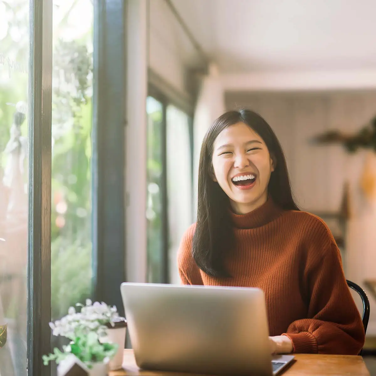 A young woman with long black hair, wearing a brown turtleneck sweater, is sitting at a table with a laptop in front of her. She is laughing and looking directly at the camera. Behind her is a large window with plants on the sill, letting in natural light. The setting appears to be a cozy, well-lit room.