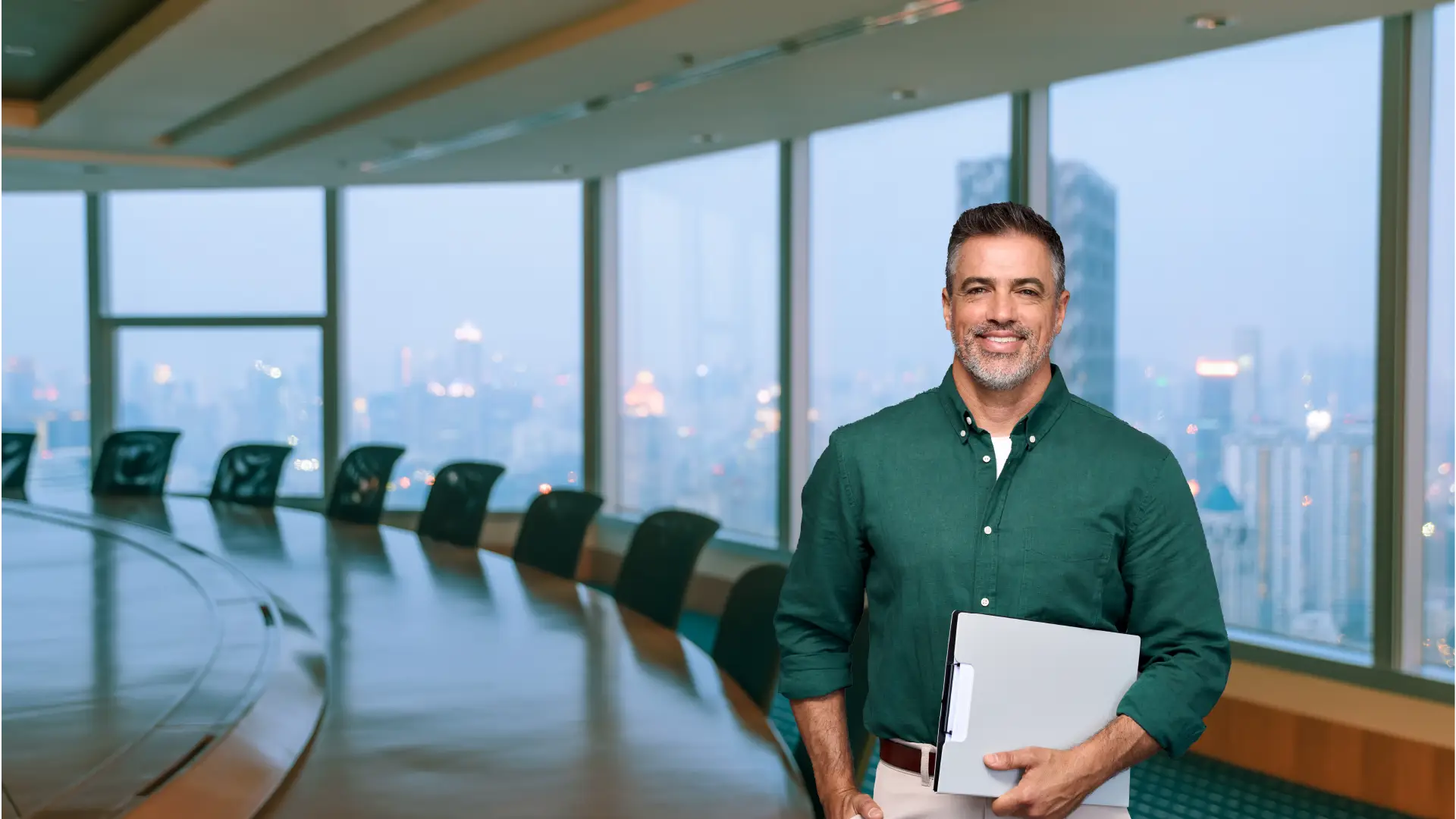 A professional man stands confidently in a modern conference room with a panoramic city view in the background. He is wearing a green button-down shirt and holding a laptop, smiling warmly at the camera. The large windows behind him provide a stunning view of a city skyline with skyscrapers and lights, adding to the sophisticated and professional atmosphere of the setting.