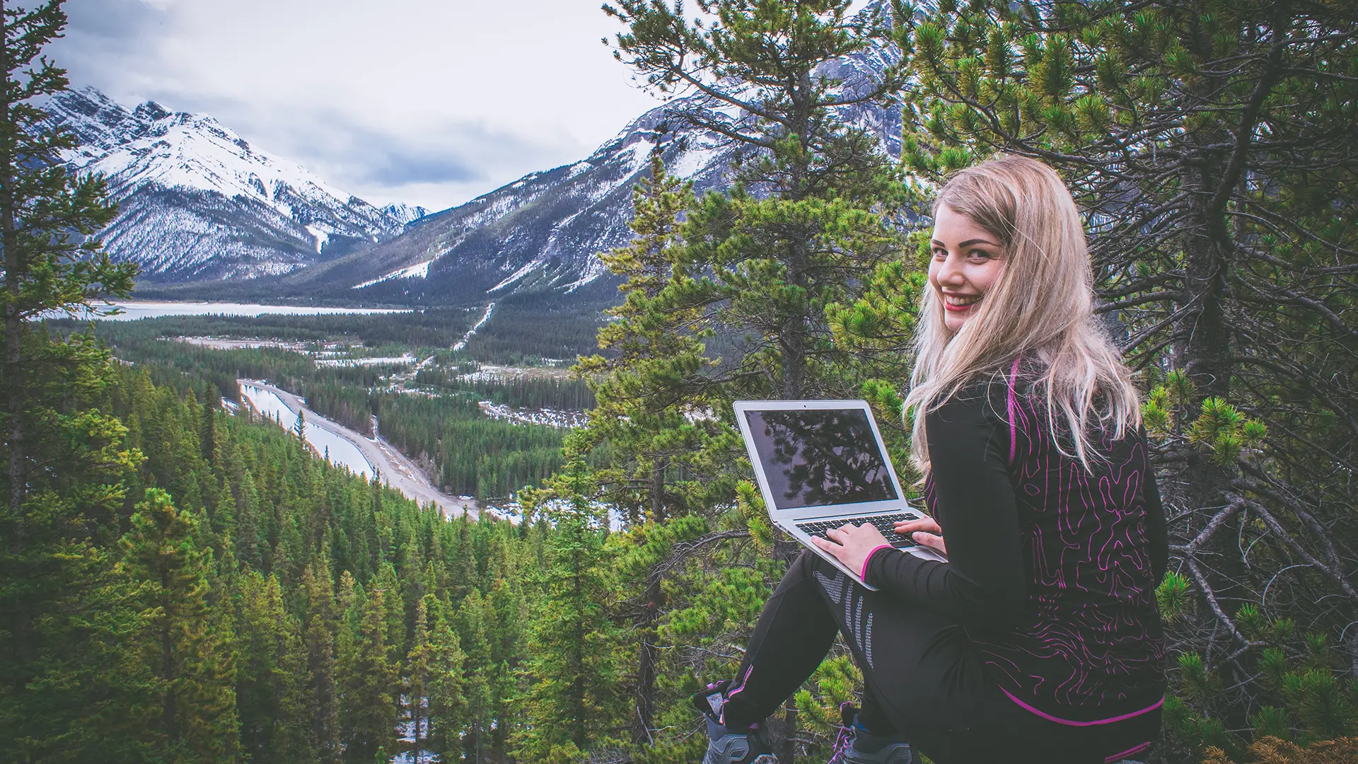 A young woman is sitting on a mountainside, working on a laptop. She is smiling and looks content, surrounded by lush green trees with a stunning view of snow-capped mountains and a winding river in the background. She is dressed in outdoor gear, indicating a love for nature and adventure.