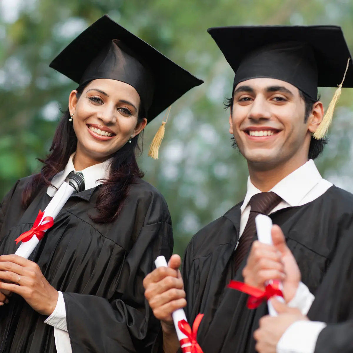 A group of international graduates stand together, smiling proudly in their black graduation gowns and caps with gold tassels. Each holds a rolled-up diploma tied with a red ribbon, signifying their academic achievement. The background is outdoors with greenery, indicating the ceremony might be taking place in a garden or park setting. This image captures the joy and accomplishment of graduation, highlighting the diversity and success of students from different backgrounds who have reached an important milestone in their educational journey.