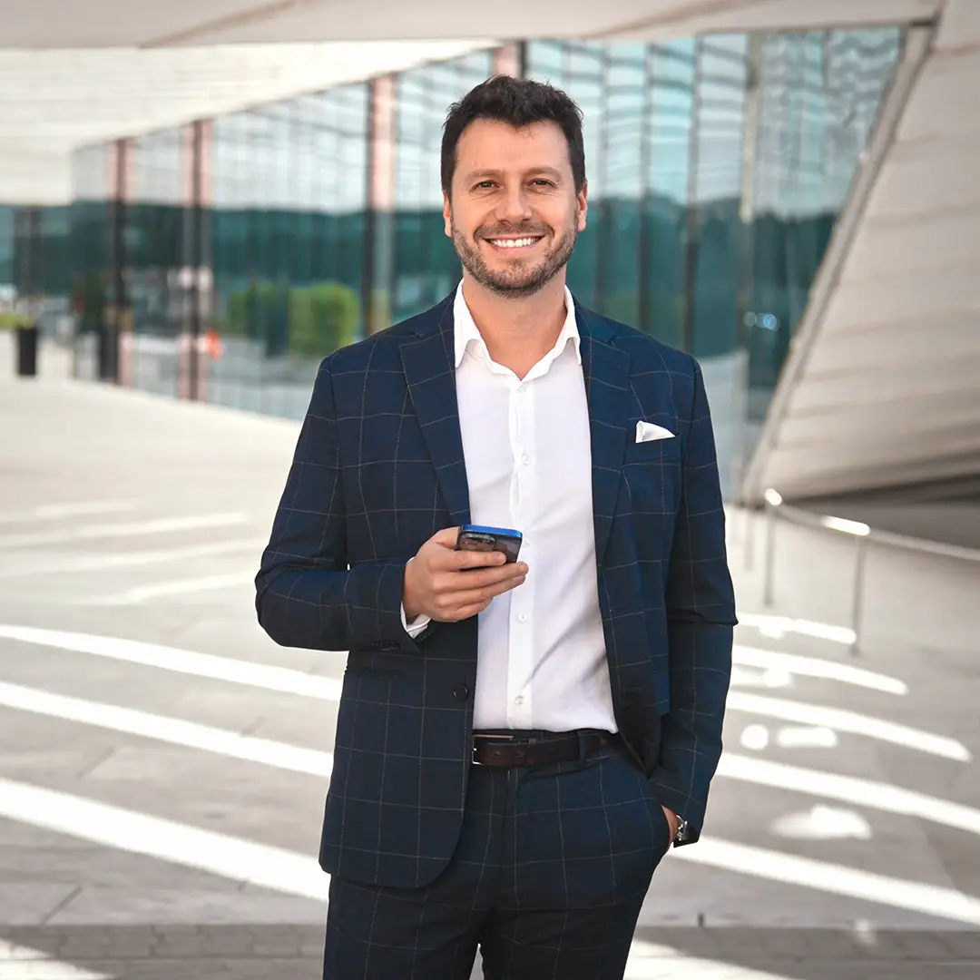 A confident professional stands outside a modern office building, smiling while holding a smartphone, symbolising leadership, connectivity, and career advancement. His polished appearance and relaxed demeanor reflect the real-world, executive-focused nature of the MBA program, designed for experienced professionals ready to take their expertise to the next level.