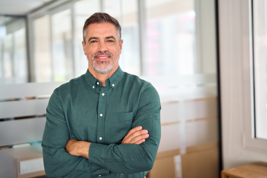 The image features a mature man with a confident and approachable demeanor, standing with arms crossed, dressed in a green shirt, and looking directly at the camera. The background appears to be an office setting with bright lighting and modern decor. This image can be used to represent "Mature leader," highlighting qualities of confidence, experience, and leadership.