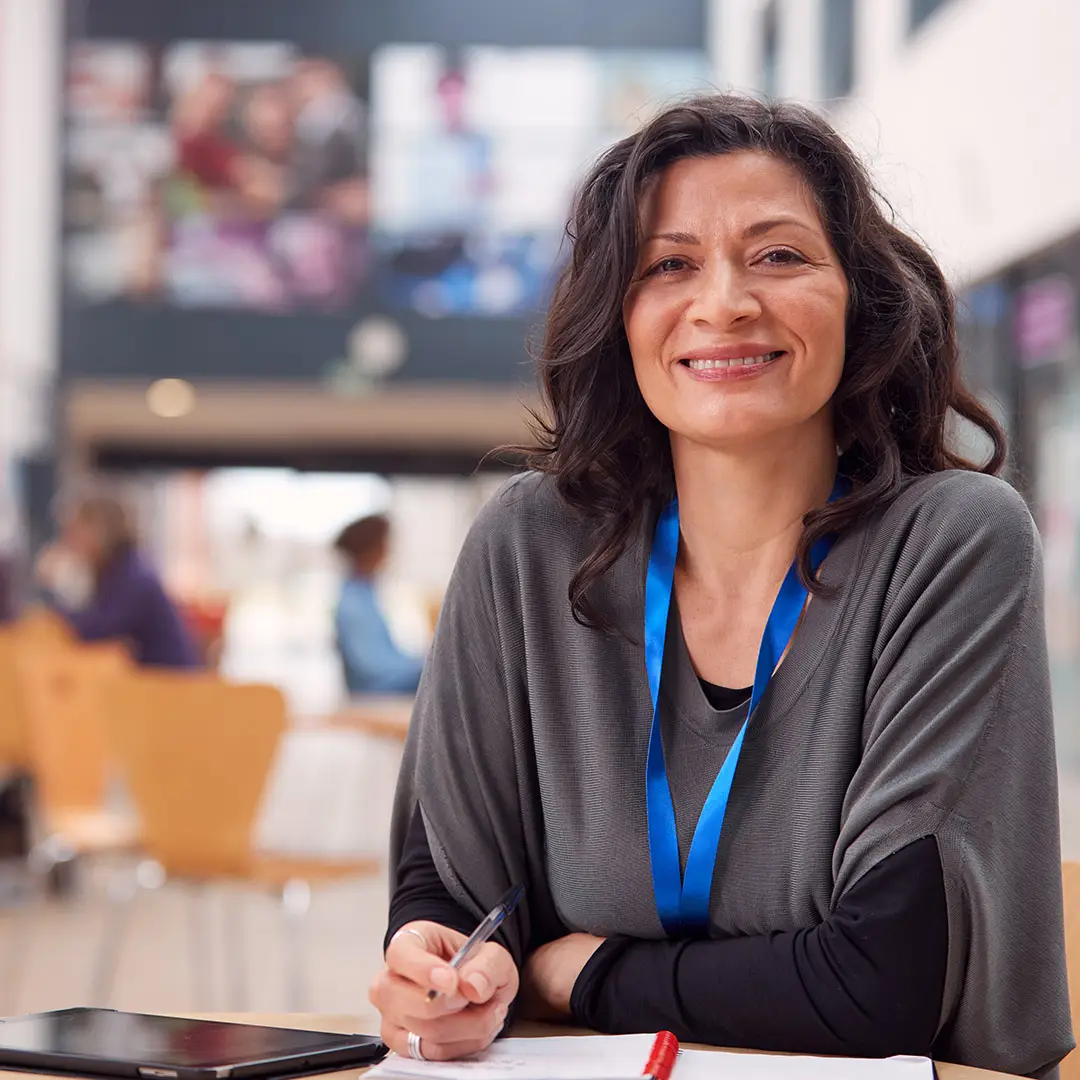 A smiling mature woman with wavy dark hair sits at a table in a modern, open learning space. She wears a blue lanyard and a grey poncho-style top over a black shirt, holding a pen over a notepad. A tablet rests on the table beside her. The blurred background shows other students in a collaborative setting, suggesting a welcoming and engaging educational environment.