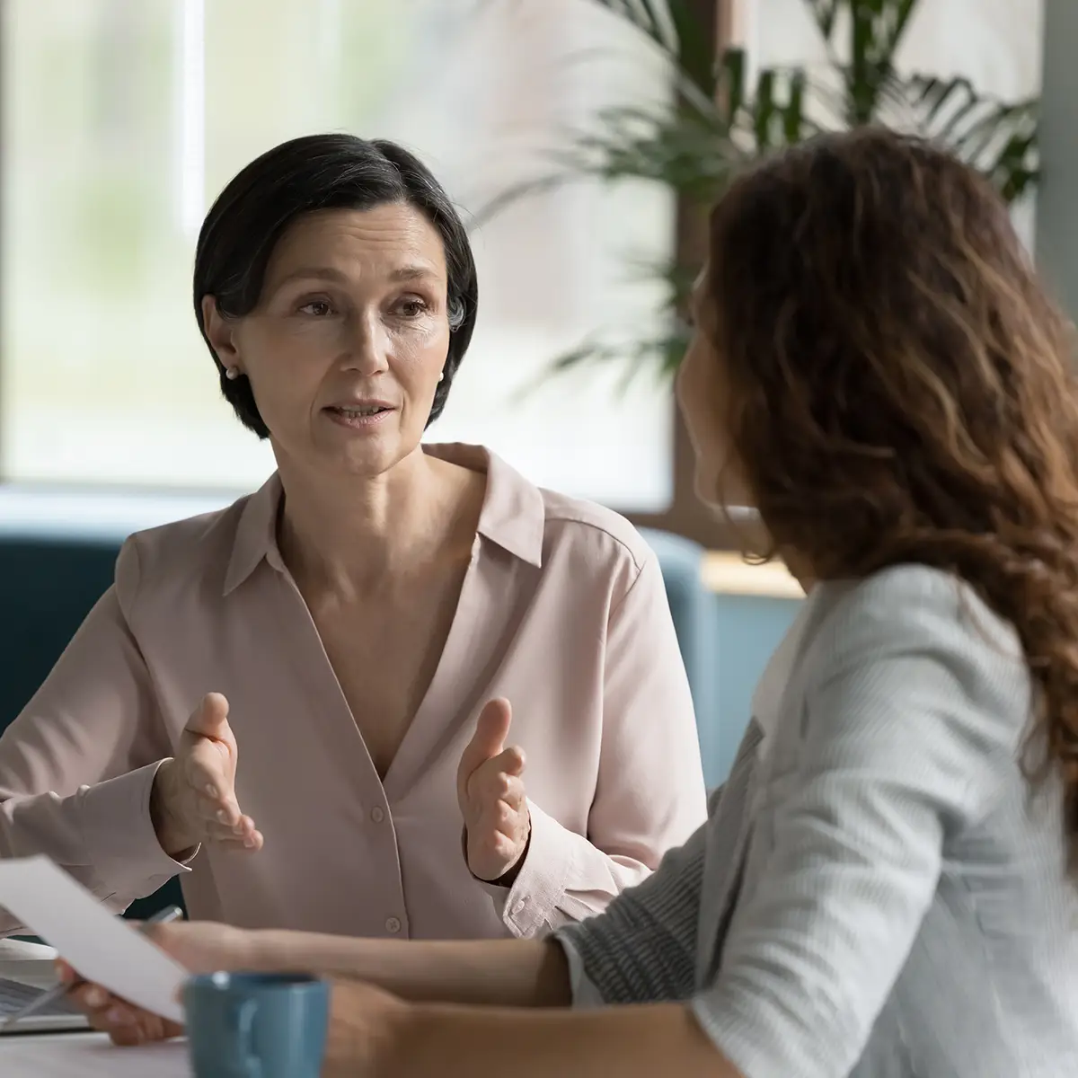 An academic mentor engages in a focused conversation with a student. The mentor gestures thoughtfully while the student listens attentively, indicating a supportive and collaborative learning environment. The setting appears to be a professional, yet comfortable space, fostering effective communication and guidance.