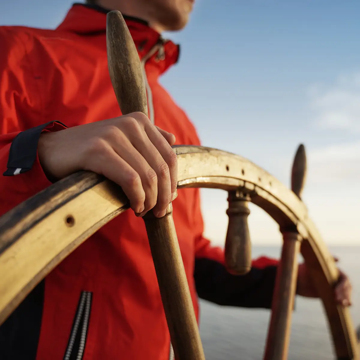 A close-up image shows a person wearing a red jacket, standing at the helm of a ship. Their hands are firmly gripping the wooden steering wheel, poised to navigate. The background features a calm sea and a clear sky, indicating a tranquil but purposeful journey. The image symbolises navigation, leadership, and the ability to steer through challenges, highlighting the importance of direction and control in achieving one's goals.