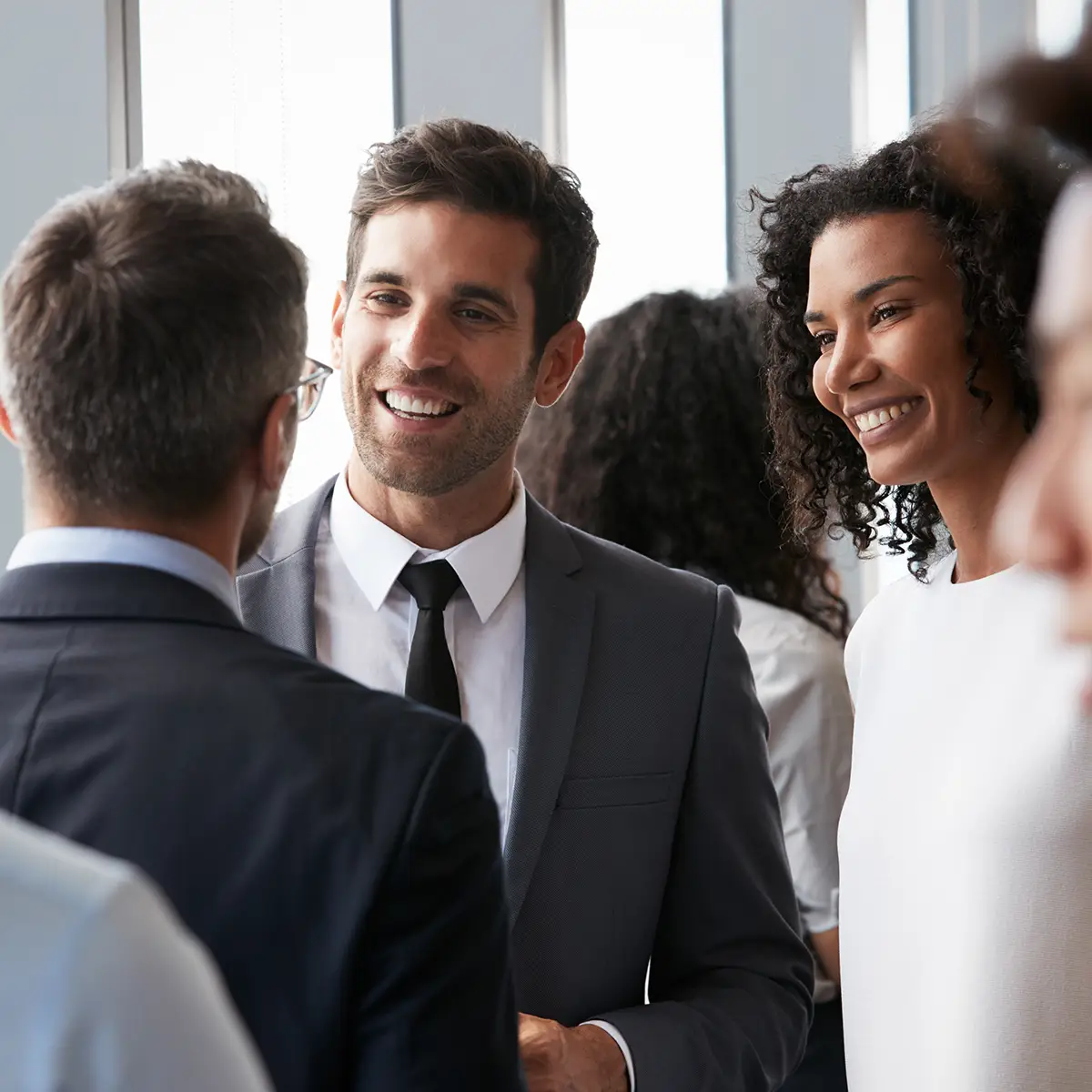 A group of professionals are engaged in a lively conversation at a networking event. The participants are dressed in business attire, and they appear to be in a modern office setting with large windows in the background. One man in a grey suit and black tie is smiling and speaking to another man in glasses, while a woman with curly hair and a white dress stands nearby, smiling warmly.