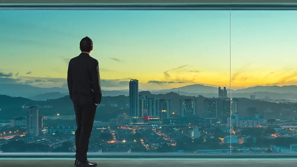 A man in a business suit stands in a large, empty room with floor-to-ceiling windows, gazing out at a panoramic cityscape at sunset. The city is bathed in the warm hues of the setting sun, with mountains in the distance and buildings illuminated by the soft evening light. The man's posture is contemplative, with his hands in his pockets, suggesting he is reflecting on his goals and aspirations. This image symbolises ambition, vision, and the pursuit of success, highlighting the importance of taking a moment to reflect on one's objectives and the broader perspective of one's journey.