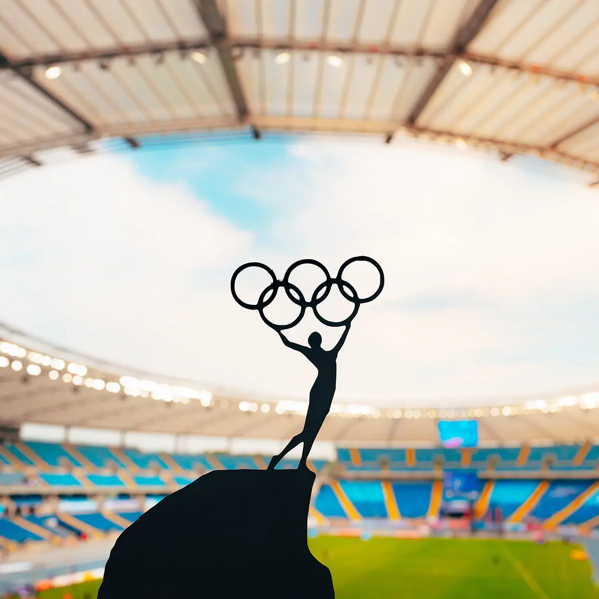 Silhouette of a figure holding the Olympic rings high above a stadium. The scene is set inside a large sports arena with rows of empty seats, capturing the iconic Olympic symbol against the backdrop of the sky and stadium roof. The image conveys a sense of achievement and global unity.