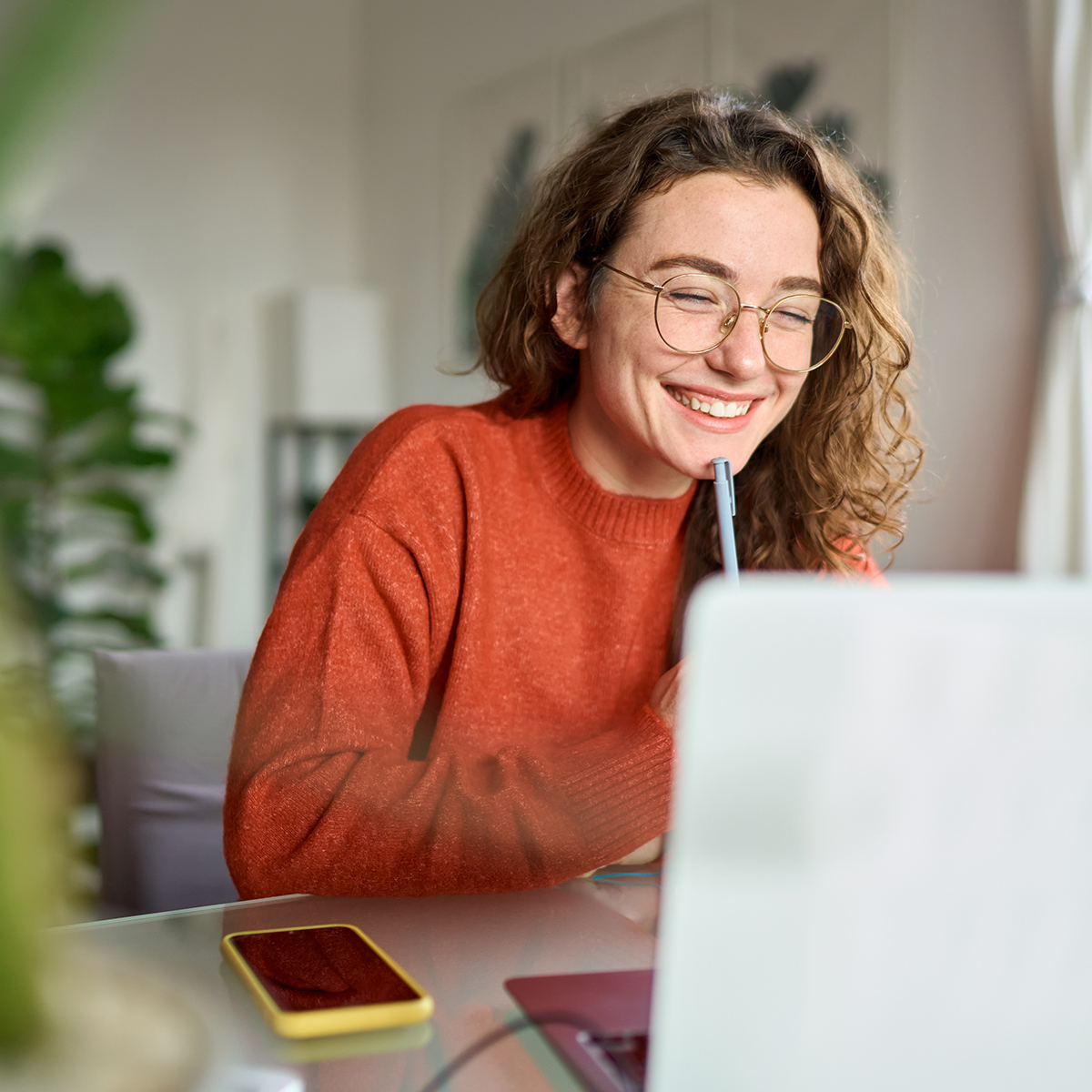 A person engaged in online learning. They are smiling, wearing glasses, and seem to be enjoying the learning experience. They have a pen in hand and are looking at a laptop, suggesting they might be attending an online class or working on a project. There is also a smartphone on the table, indicating they might be using multiple devices for their learning activities. The setting is bright and appears to be a comfortable, home-like environment.