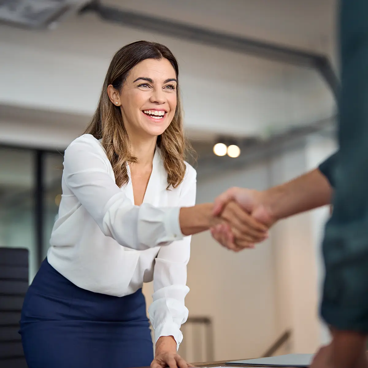 A professional woman in a business setting smiles warmly as she shakes hands with a colleague, symbolising academic and industry partnerships. Her confident and friendly demeanour reflects the positive collaboration between academic institutions and industry professionals, fostering mutual growth and innovation. The modern office environment underscores the dynamic and productive nature of these partnerships.