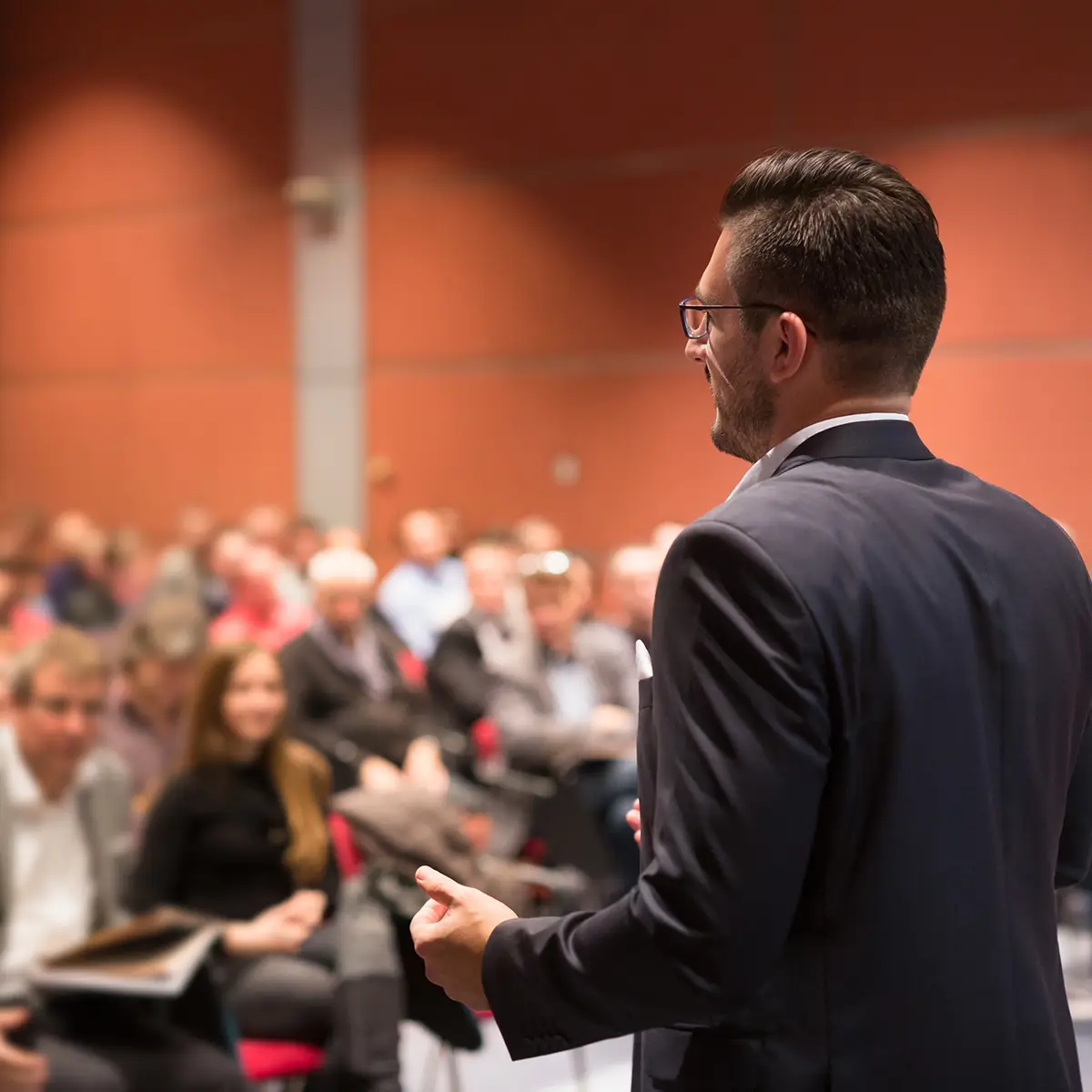 A business expert, who is also a professor, speaks confidently to a large audience in a lecture hall. The professional is dressed in a suit, engaging the attendees with his insights, highlighting his dual role in academia and industry. The attentive audience underscores the value of his expertise, blending practical business knowledge with academic teaching.