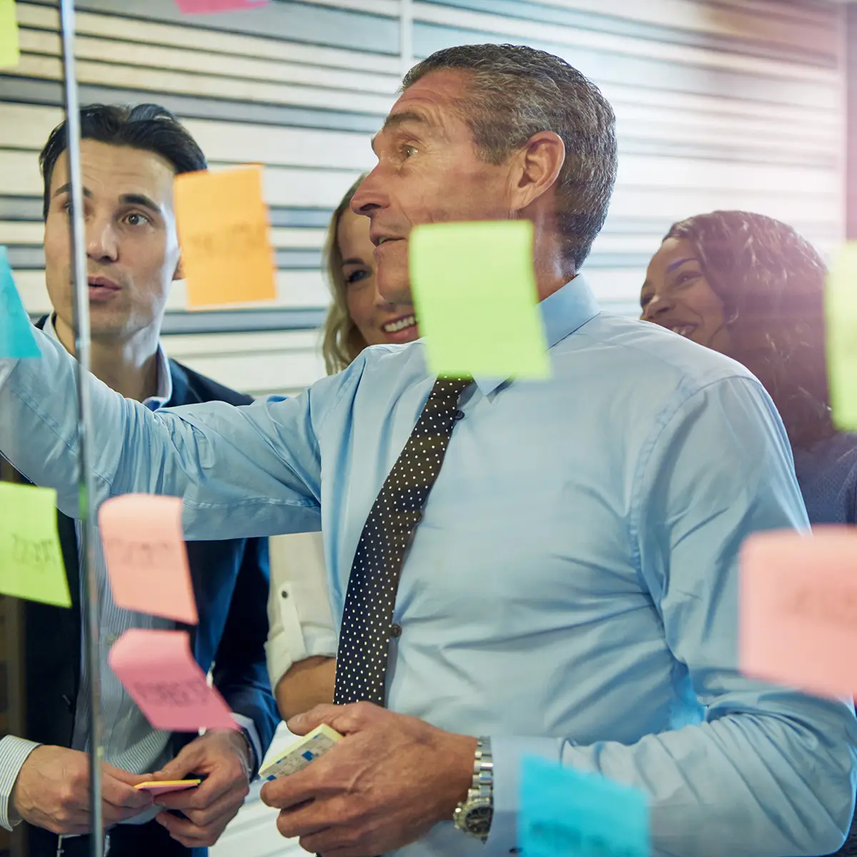 A team collaborates enthusiastically in a project-based learning environment, using colourful sticky notes on a glass wall to organise their ideas. The central figure, an experienced leader in a blue shirt and polka dot tie, gestures towards the notes, guiding the discussion. The diverse group of participants listens attentively and contributes actively, illustrating the dynamic and interactive nature of project-based learning.