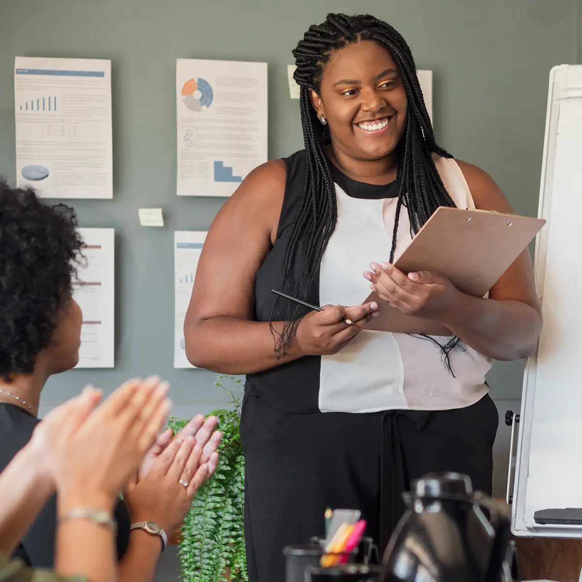 A confident African-American woman stands in front of a group, holding a clipboard and smiling as she engages with her audience. She is dressed in a stylish, professional outfit and appears to be leading a presentation or discussion. Behind her, a wall displays various charts and documents, adding a sense of professionalism and purpose to the setting. The audience members are clapping, indicating appreciation and support.