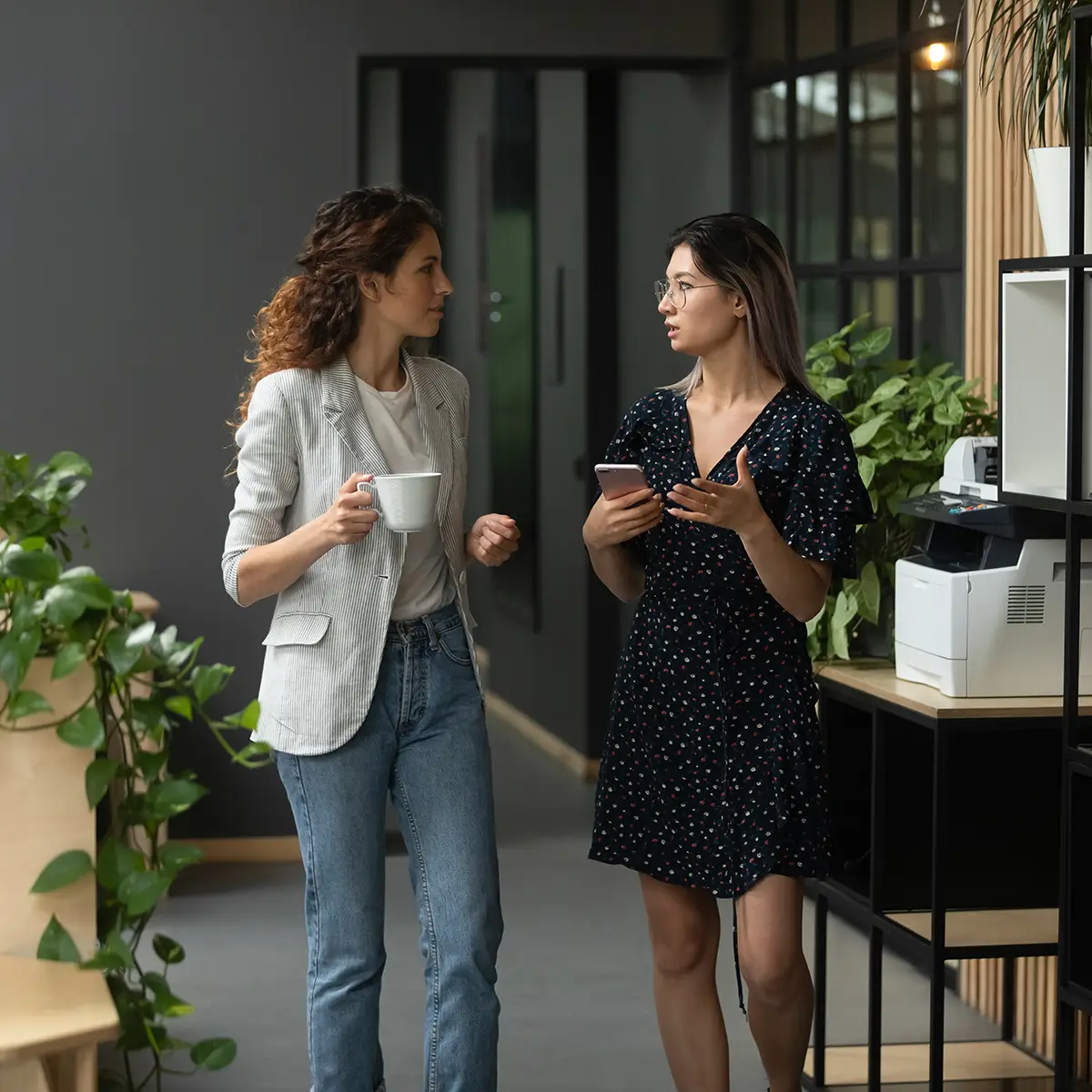 Two women are engaged in a conversation in a modern office setting. One woman, dressed casually in jeans and a blazer, holds a coffee mug, while the other woman, in a patterned dress, holds a smartphone and gestures as she speaks. They are walking together through a hallway adorned with greenery and modern office equipment. The scene suggests one woman is showing the other around, providing guidance or sharing information about the workplace. This image captures the concept of "showing the ropes," highlighting mentorship, support, and the process of helping someone acclimate to a new environment.