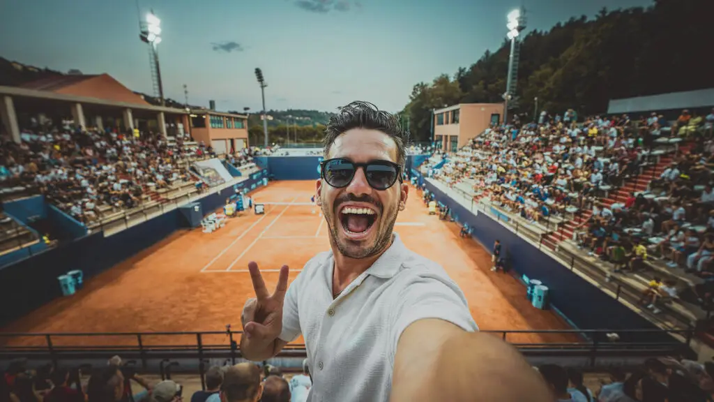 Excited sports fan taking a selfie at a tennis match, smiling and flashing a peace sign. The clay court and full stadium create a lively atmosphere, with the match in progress in the background under stadium lights. The fan is wearing sunglasses, capturing the energy of the event.