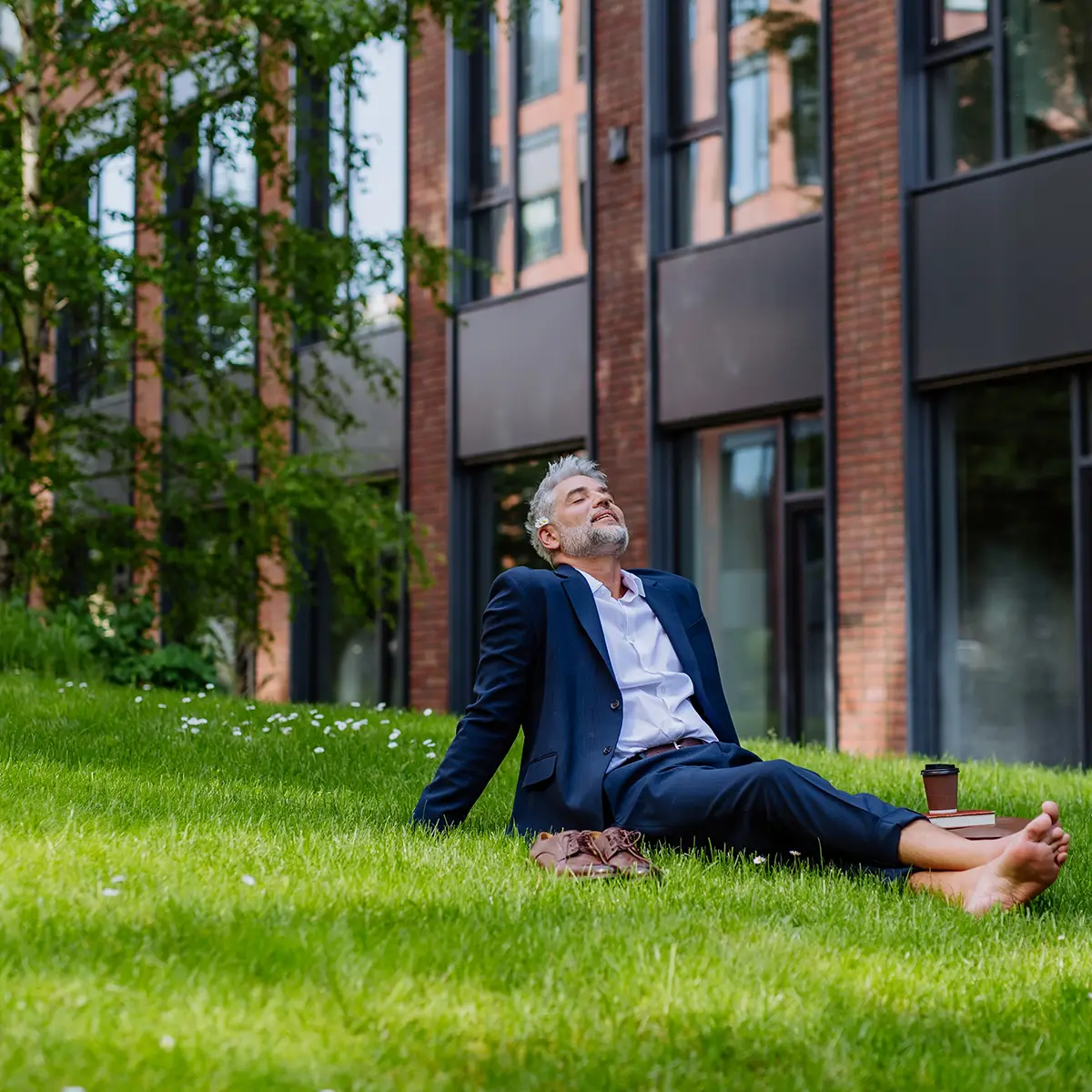 A person in a suit relaxes barefoot on a grassy lawn outside a modern building, eyes closed and face tilted towards the sun, embodying a peaceful work/study/life balance. Nearby, a coffee cup and a book are placed on the grass, suggesting a break from work or study to enjoy the outdoors.