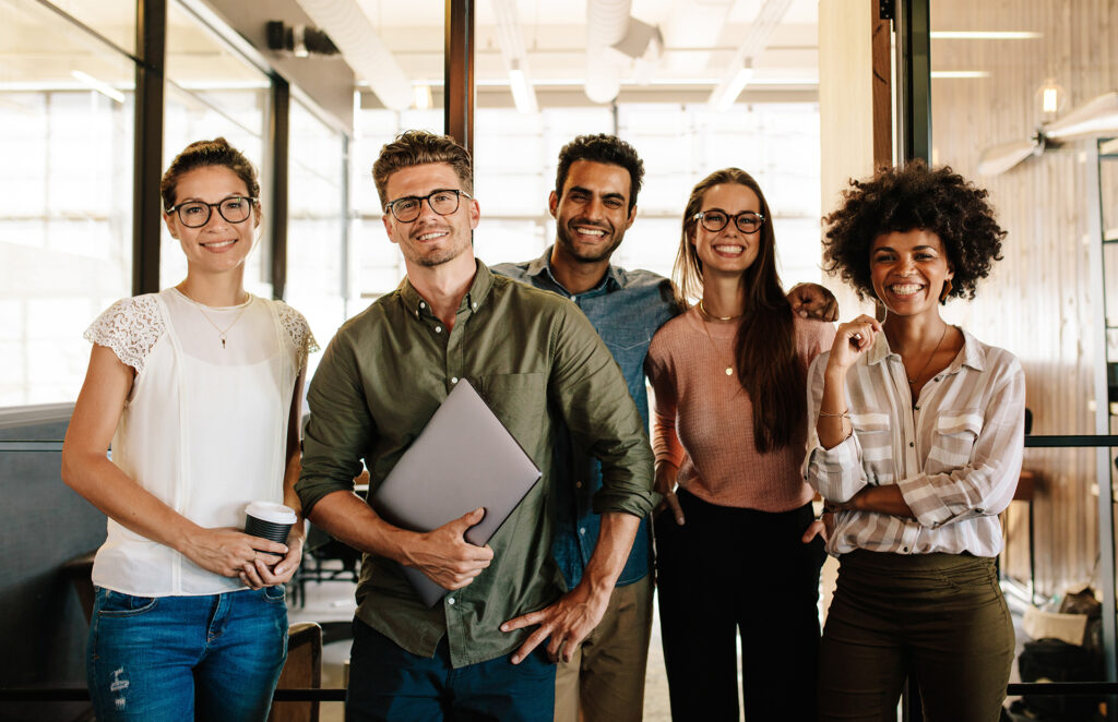 The image shows a diverse group of young professionals standing together, smiling, and looking confident. Each person is dressed in casual business attire, and they appear to be in a modern office or co-working space. This image can be used to represent "Successful students" in a university or educational setting, highlighting diversity, collaboration, and a positive, forward-thinking atmosphere.