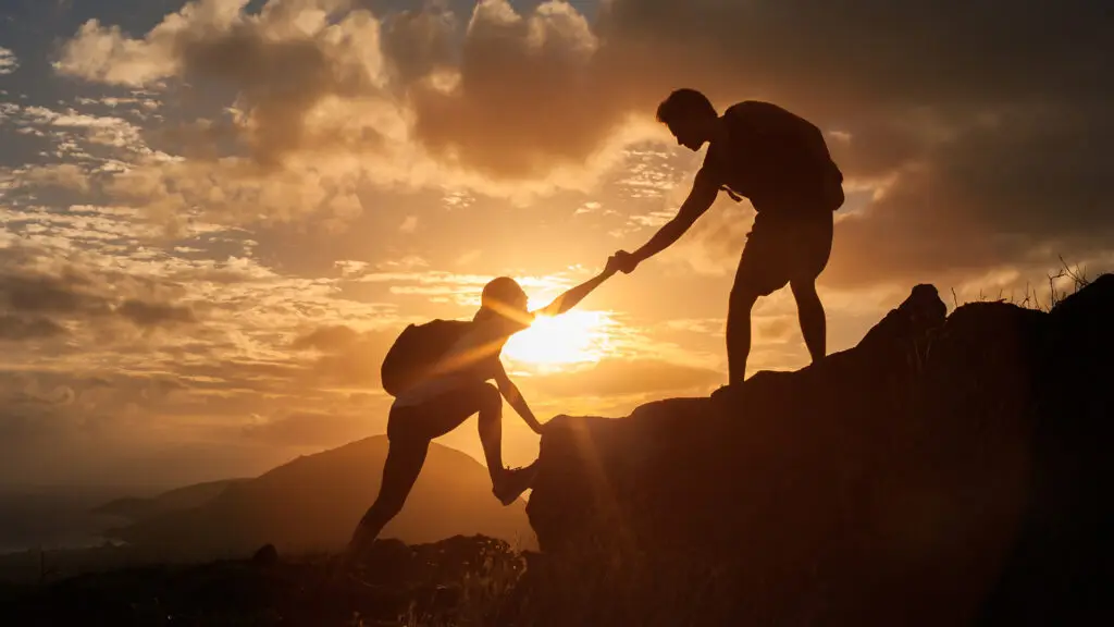 Two individuals silhouetted against a stunning sunset, with one reaching out to help the other climb a rocky hill, symbolising personalised support. The image captures the essence of guidance and assistance, emphasising the importance of one-on-one help in overcoming challenges and achieving goals. The natural, serene setting enhances the theme of supportive relationships and mutual aid.