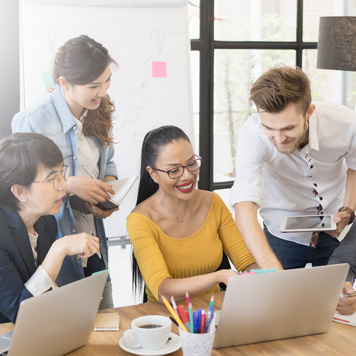 The image shows a diverse group of professionals engaged in a collaborative session, likely working on a project or training module. The group is gathered around a table with laptops, notebooks, and a whiteboard in the background filled with diagrams and post-it notes. This setting is ideal for representing "Tailored Training," highlighting the personalised and interactive approach to professional development where individuals can work together to enhance their skills and knowledge.