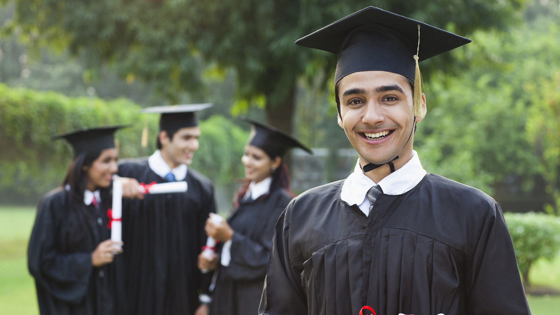 The image shows a group of young graduates in their caps and gowns, celebrating their achievement. One graduate in the foreground is smiling brightly, holding a diploma. The background features other graduates in conversation, holding their diplomas as well. This image is ideal for representing "Undergraduate Programs," showcasing the joy and accomplishment of completing a degree.