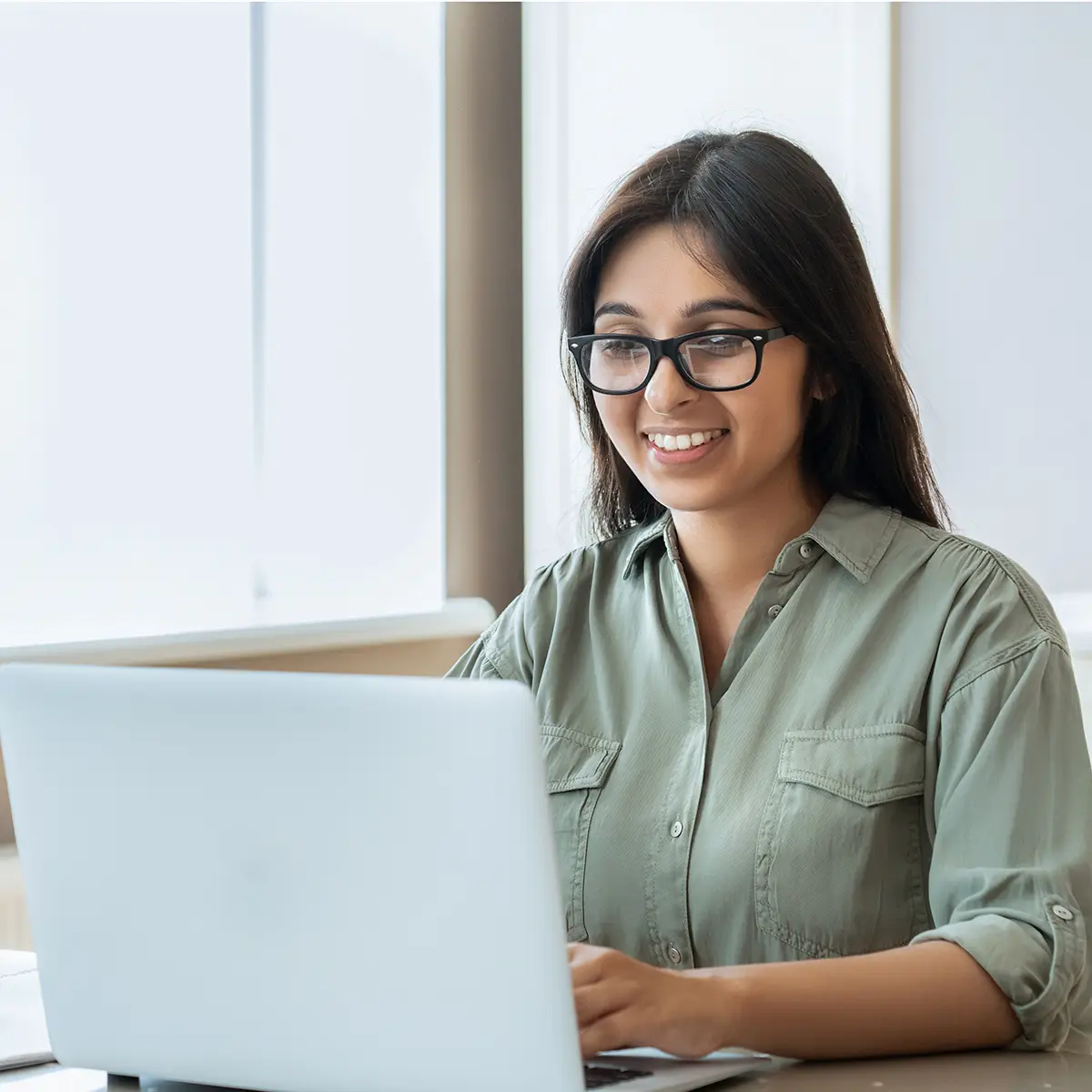 A young woman wearing glasses and a green shirt sits at a desk, smiling as she works on her laptop. The room is bright and filled with natural light from a large window behind her. A potted plant adds a touch of greenery to the workspace, creating a pleasant and inviting atmosphere. This image captures the essence of a focused and positive work or study environment, emphasising productivity, engagement, and the comfort of working in a well-lit and thoughtfully arranged space.